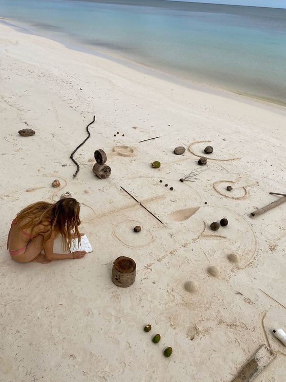 Person writing in a notebook on a beach with shells and driftwood arranged on the sand.